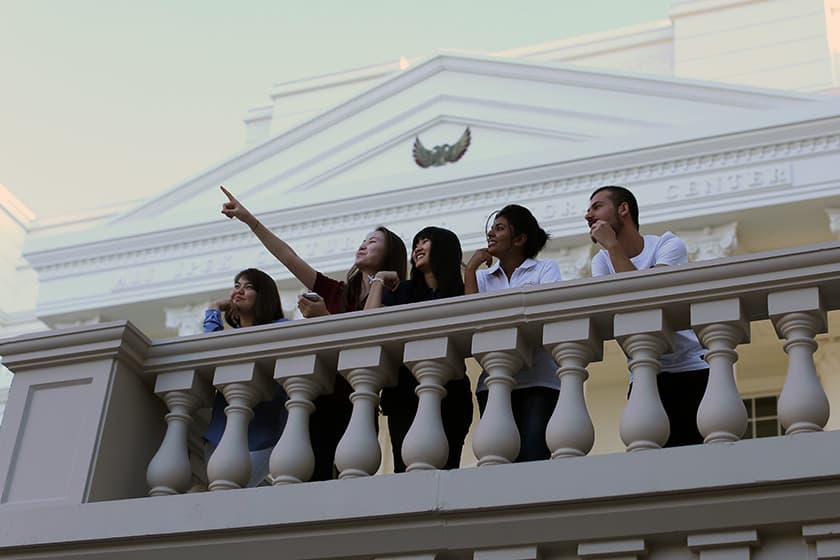 Five students standing together on a white stone campus balcony, with a pedimented building behind them.