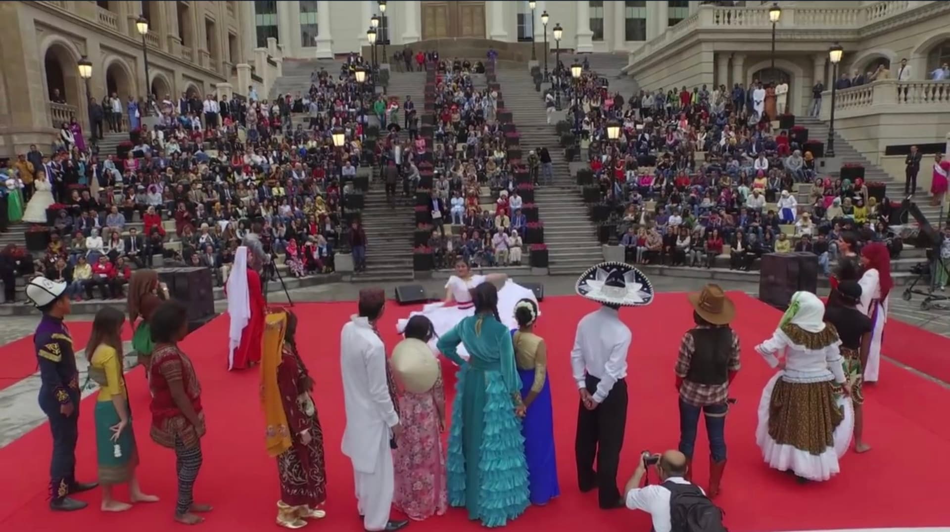 Students in the traditional dress of many countries standing on a red carpet before the campus’s grand stairs, watched by a crowd filling the steps.