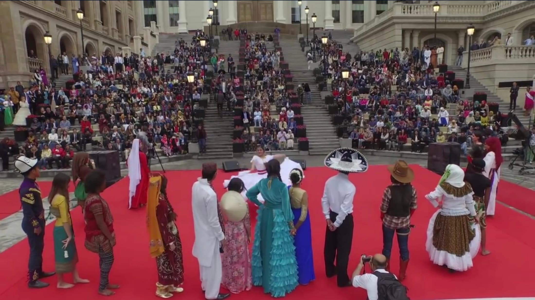 Students in the traditional dress of many countries standing on a red carpet before the campus’s grand stairs, watched by a crowd filling the steps.