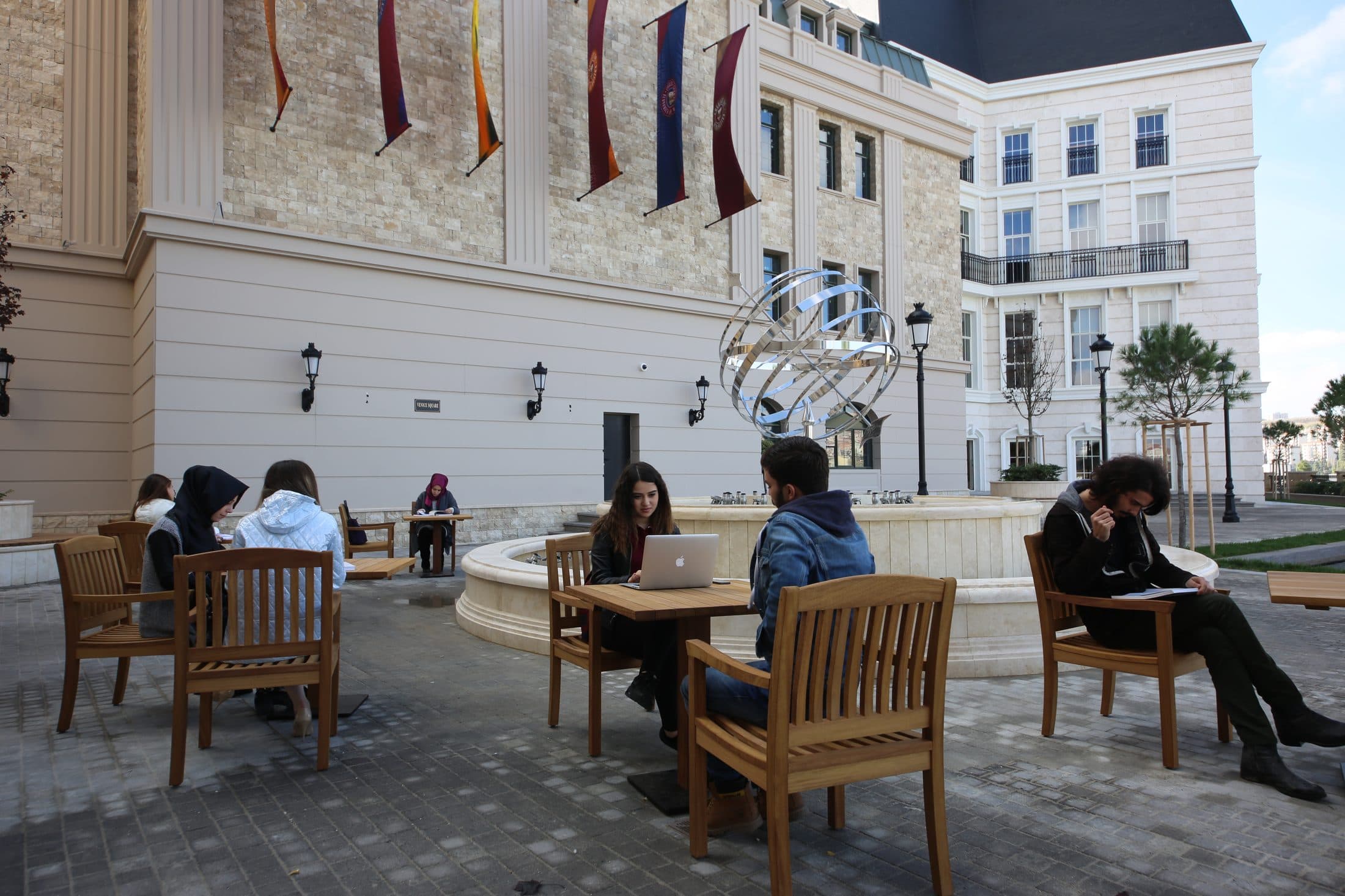 Students seated at outdoor tables in a campus square, with a stone fountain and a metal sphere sculpture at the centre.