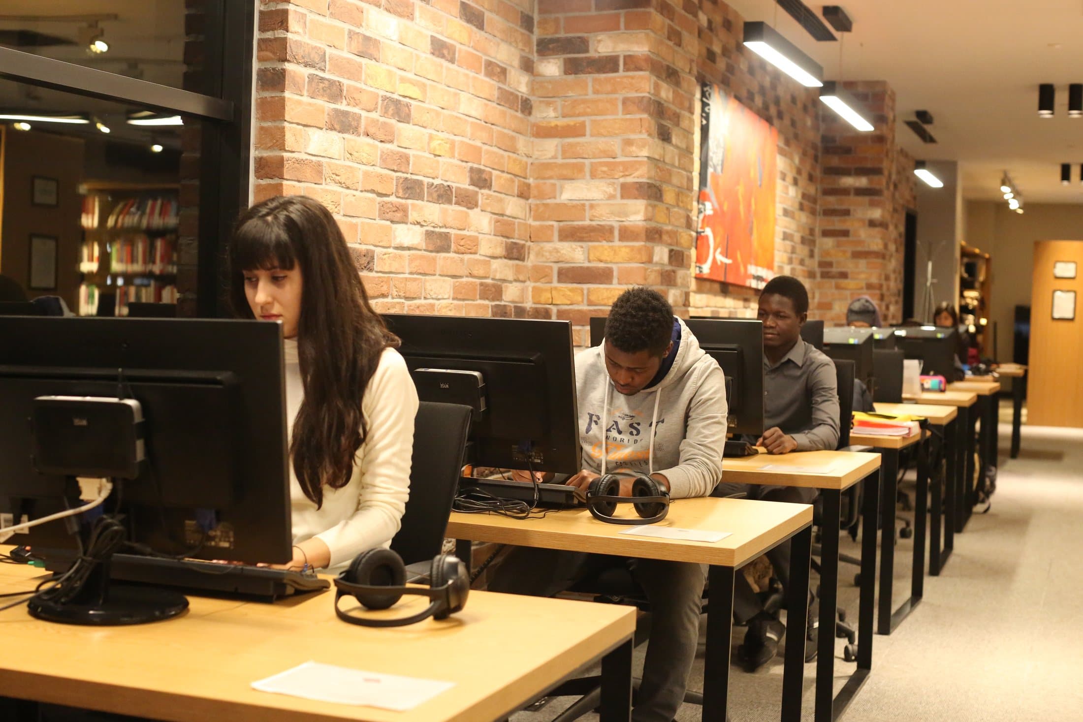 Students working at computer desks; bookshelves are visible beyond the glass.