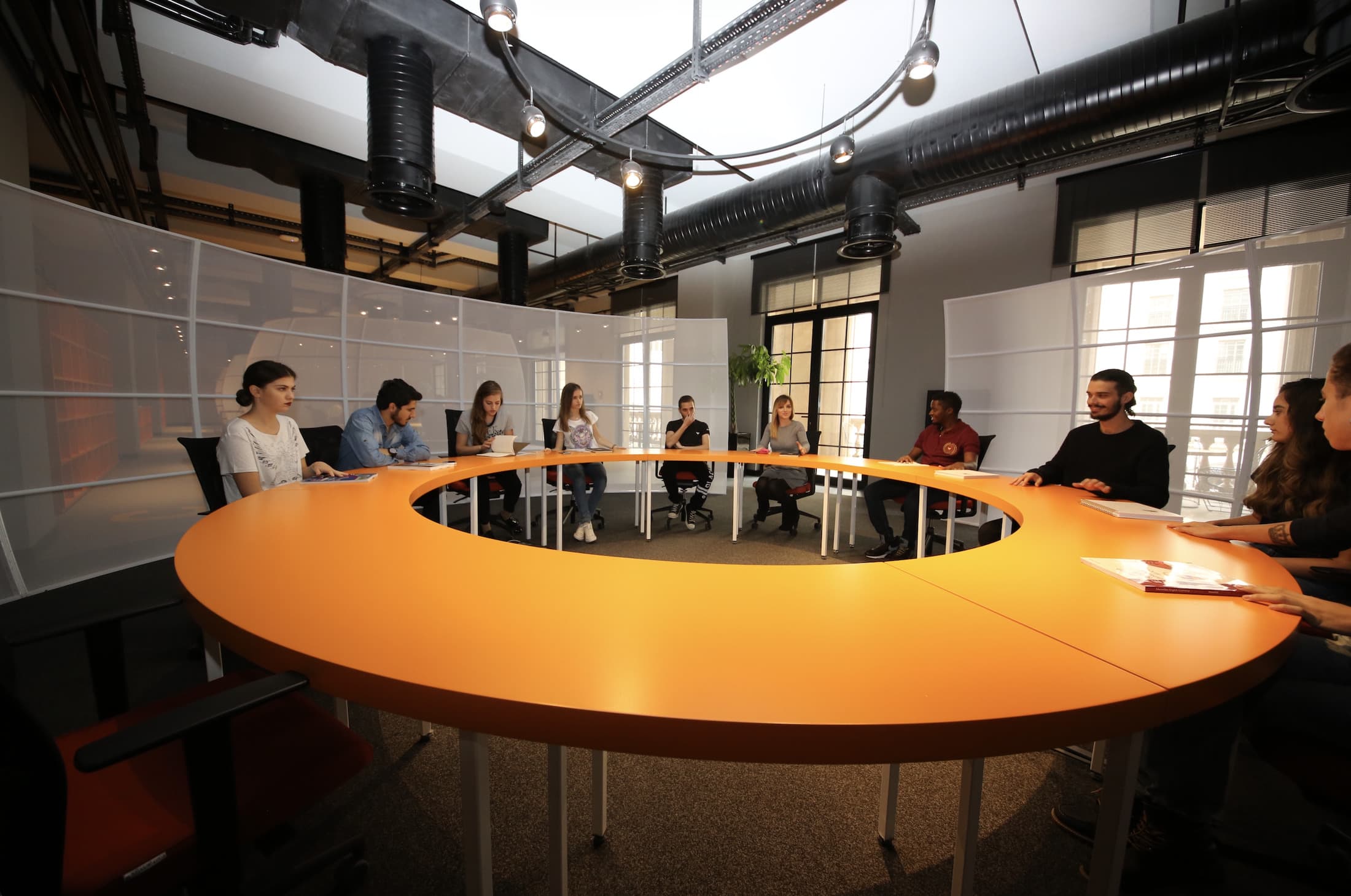 Students seated around an orange ring table in a workspace with an industrial ceiling.